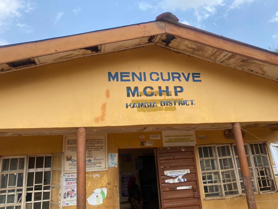 Yellow building with a sign that reads 'Menu Curve M.C.H.P Kambia District' and rusty support poles in front of the entrance, windows on both sides, and a person visible inside.