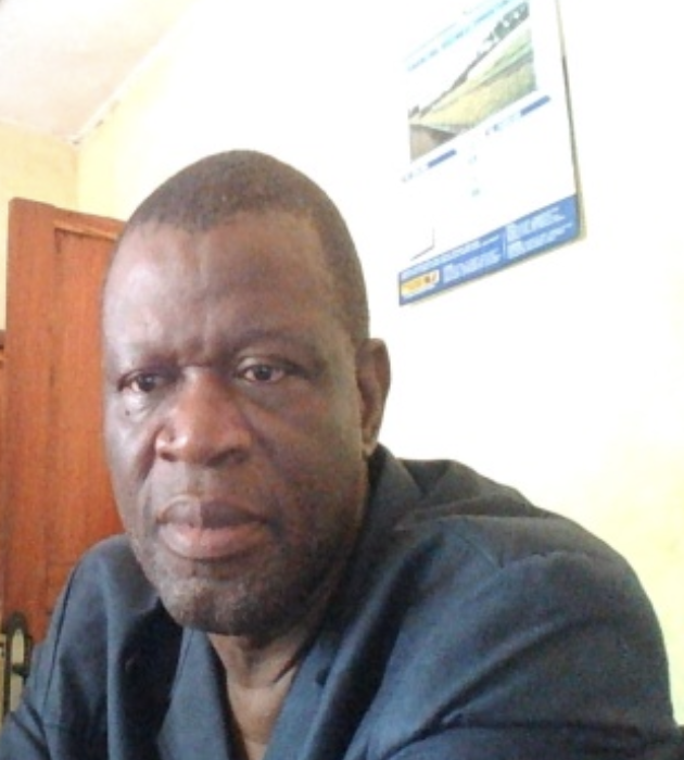 A middle-aged man with short hair and a beard, wearing a dark jacket, looking into the camera in an indoor setting with a wooden door and a calendar on the wall behind him.