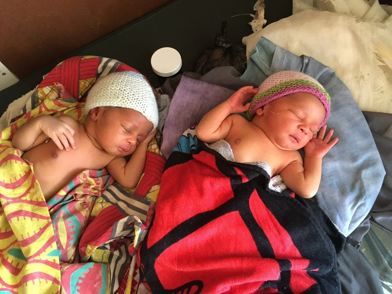 Two newborn babies sleeping on a bed, each wrapped in a blanket and wearing a knit hat.