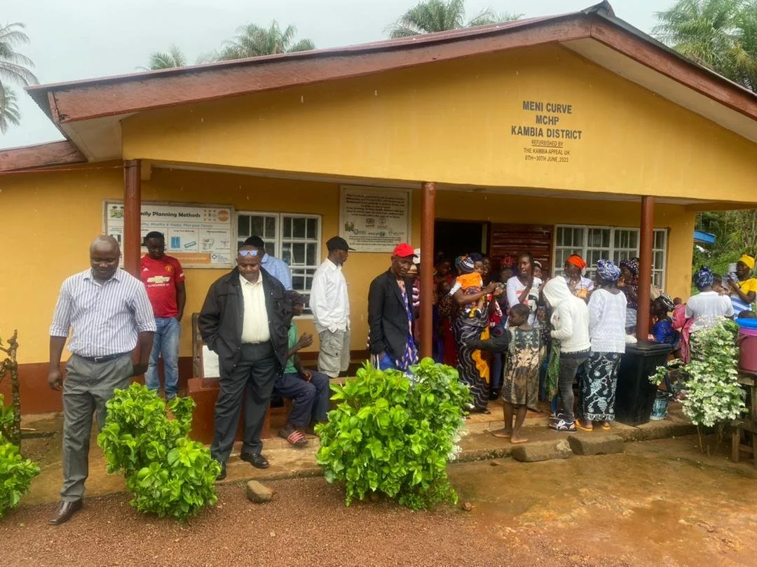 Group of people standing outside a yellow community building with a sign stating 'Meni Curve MCHP Kambia District.' Many women wearing colorful headscarves and men are gathered near the entrance.