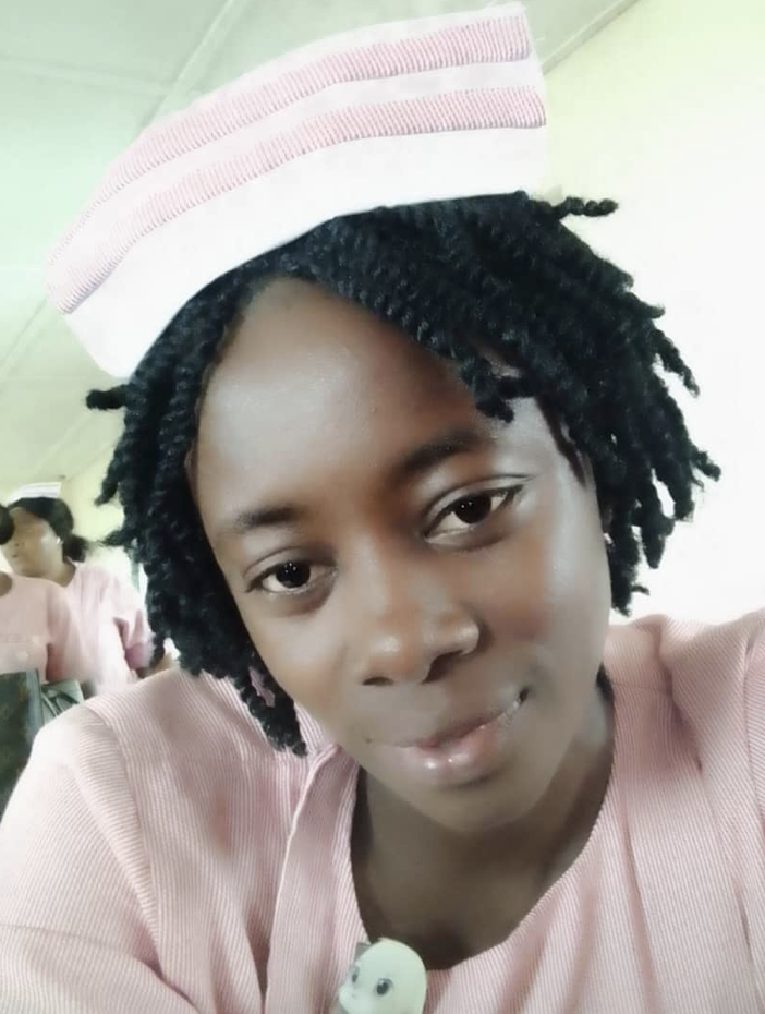 A nurse in pink scrubs and a striped cap is taking a selfie in a healthcare setting. She has short, textured black hair and is smiling softly. There are other healthcare workers in pink scrubs in the background.