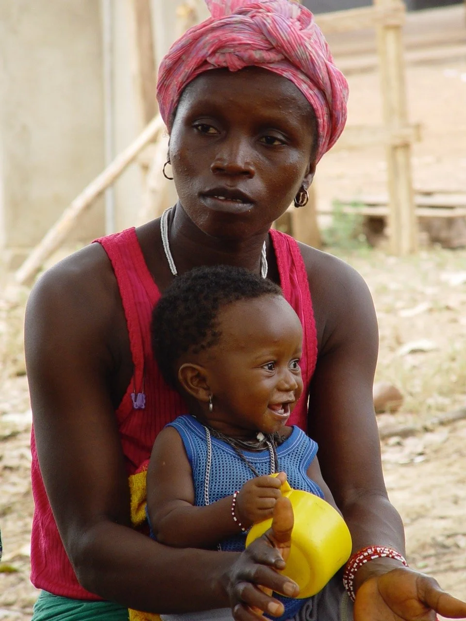 A woman wearing a pink headscarf and red top is holding a smiling young child with short, curly hair, dressed in a blue outfit, outdoors with a wooden structure in the background.