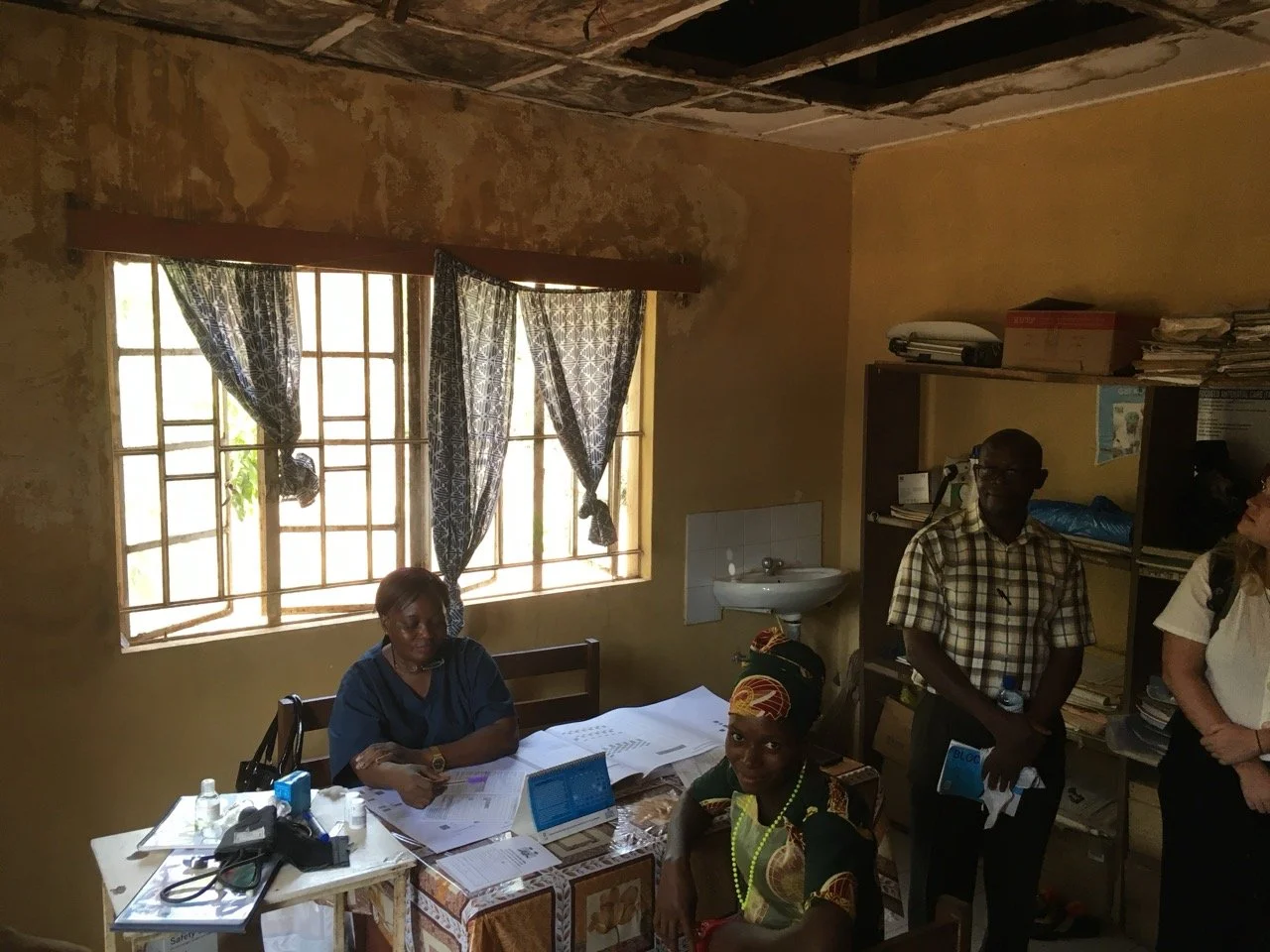 Four people in a small office with a window and shelves. One woman is seated at a desk with papers and a laptop, another woman and two men are standing nearby. The office has a worn appearance with a partially damaged ceiling and a small sink.
