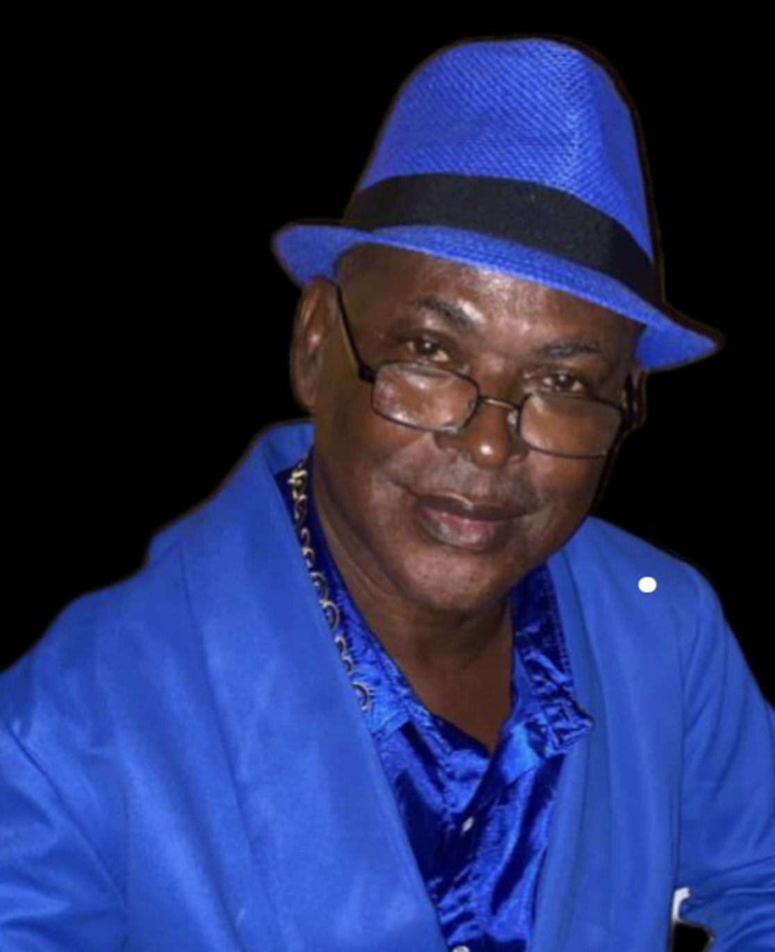 A man wearing a bright blue fedora hat, glasses, a blue suit, and a patterned shirt, smiling at the camera against a black background.