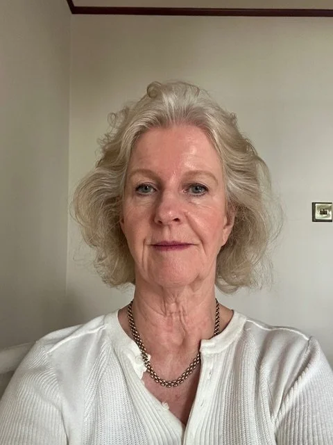A close-up of an older woman with shoulder-length curly gray hair, wearing a white top and a silver necklace, standing against a plain background.