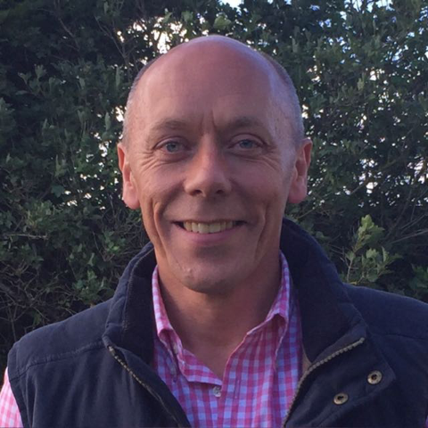 A smiling man with a shaved head, wearing a pink checkered shirt and a dark jacket, standing outdoors in front of green bushes.