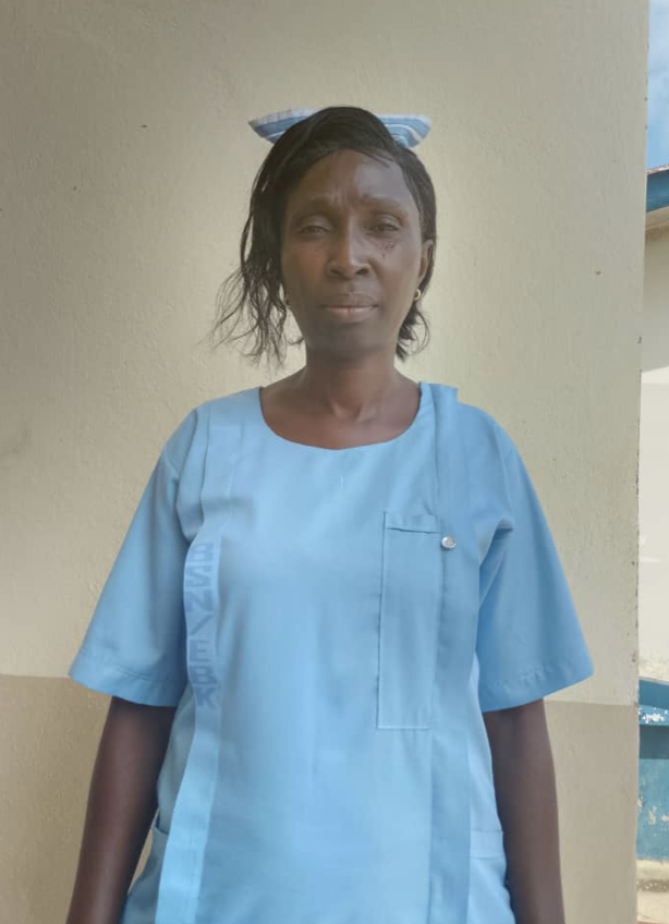 A woman dressed in a light blue healthcare uniform standing outdoors against a beige wall. She has short, dark hair and a neutral expression.