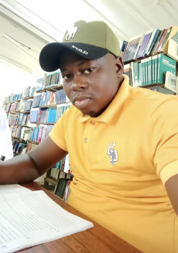 A young man in a yellow polo shirt and a baseball cap sitting at a table with a book open in front of him in a library with bookshelves in the background.
