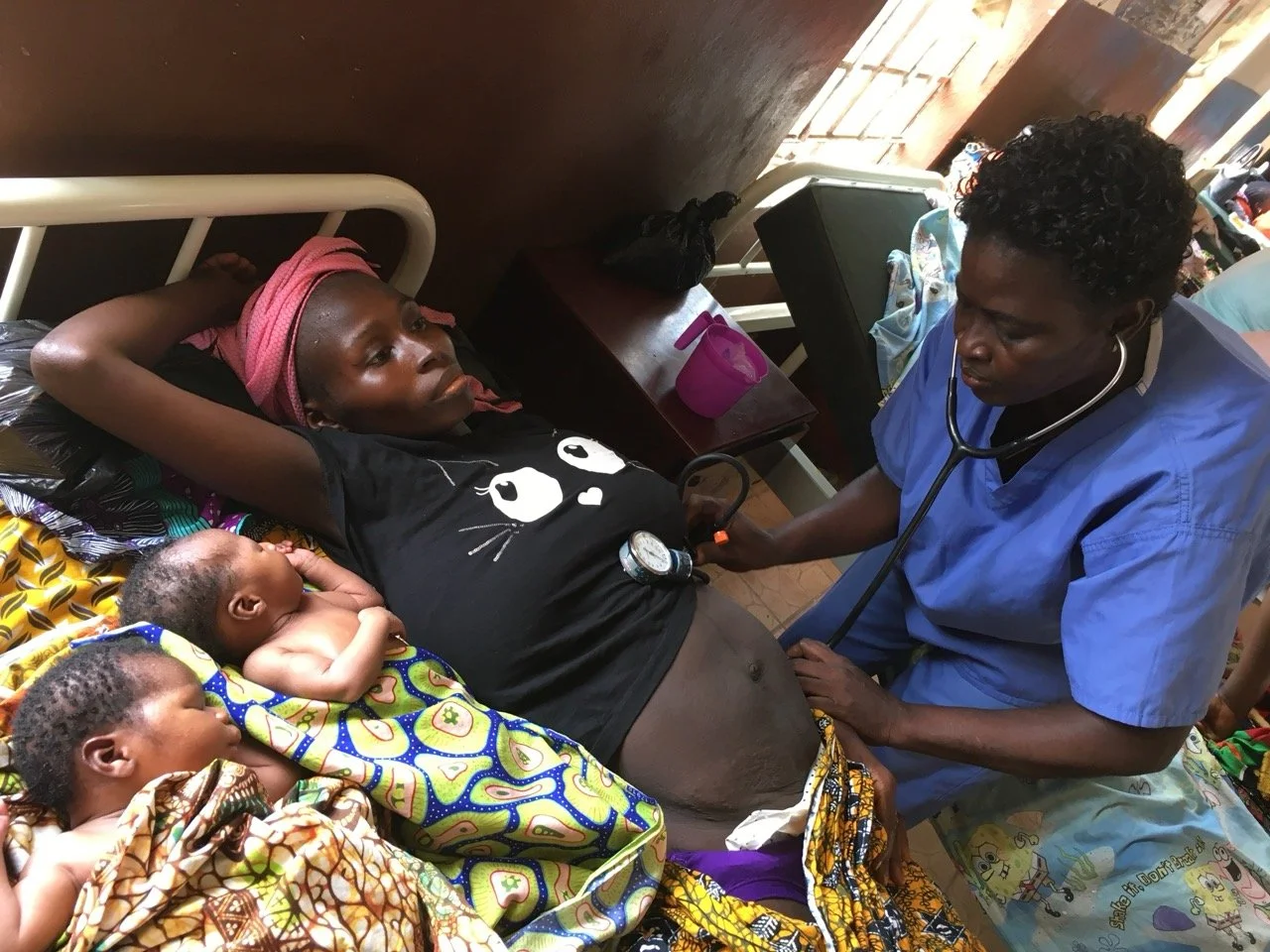 A healthcare worker using a stethoscope to check a pregnant woman lying on a bed, surrounded by two babies, in a modest room with a dark wall, a wooden table, and patterned cloths.