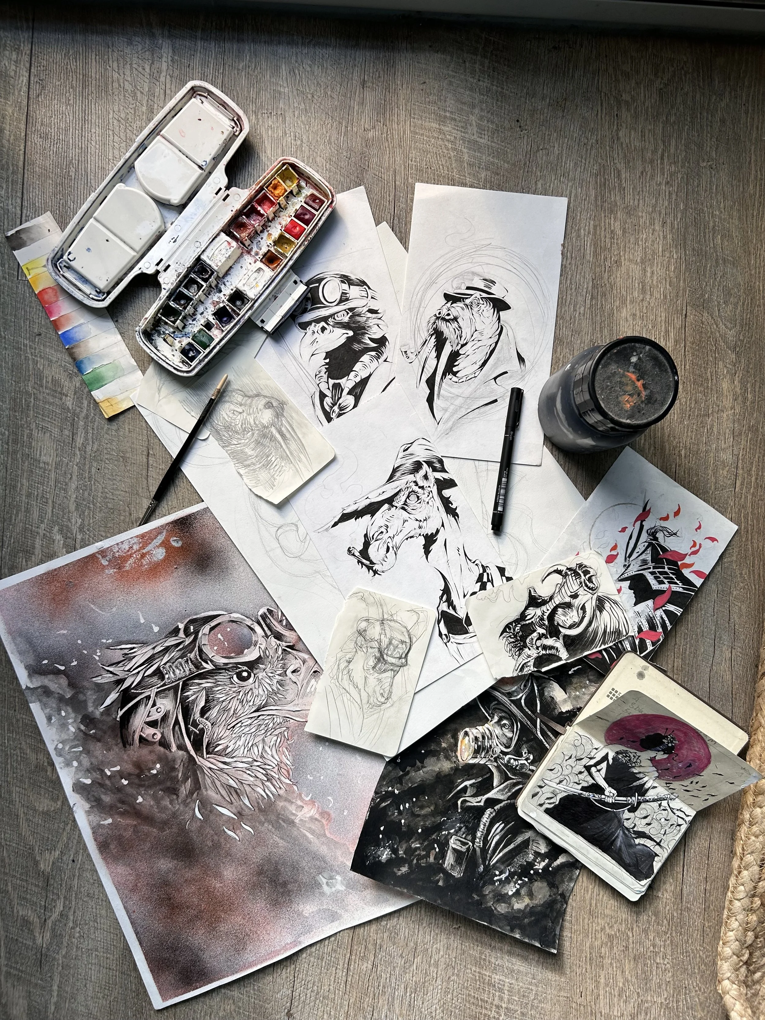 Table à terre avec des dessins sur papier, une palette d'aquarelle, un pinceau, une bouteille de liquide noir, et un jeu de cartes avec une illustration d'un personnage.