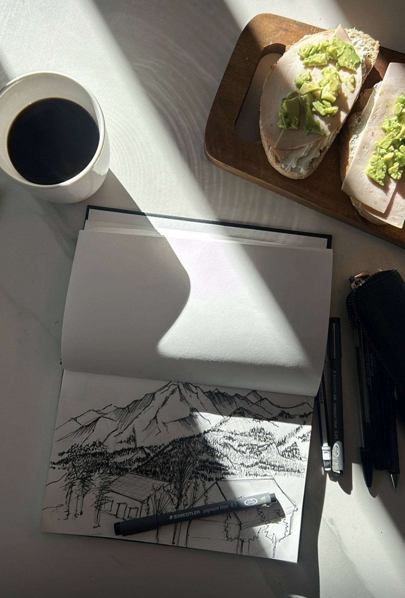 Un dessin d'une montagne avec un village au premier plan, réalisé à l'aide de crayon noir, placé sur une table avec des marqueurs et un carnet ouvert.