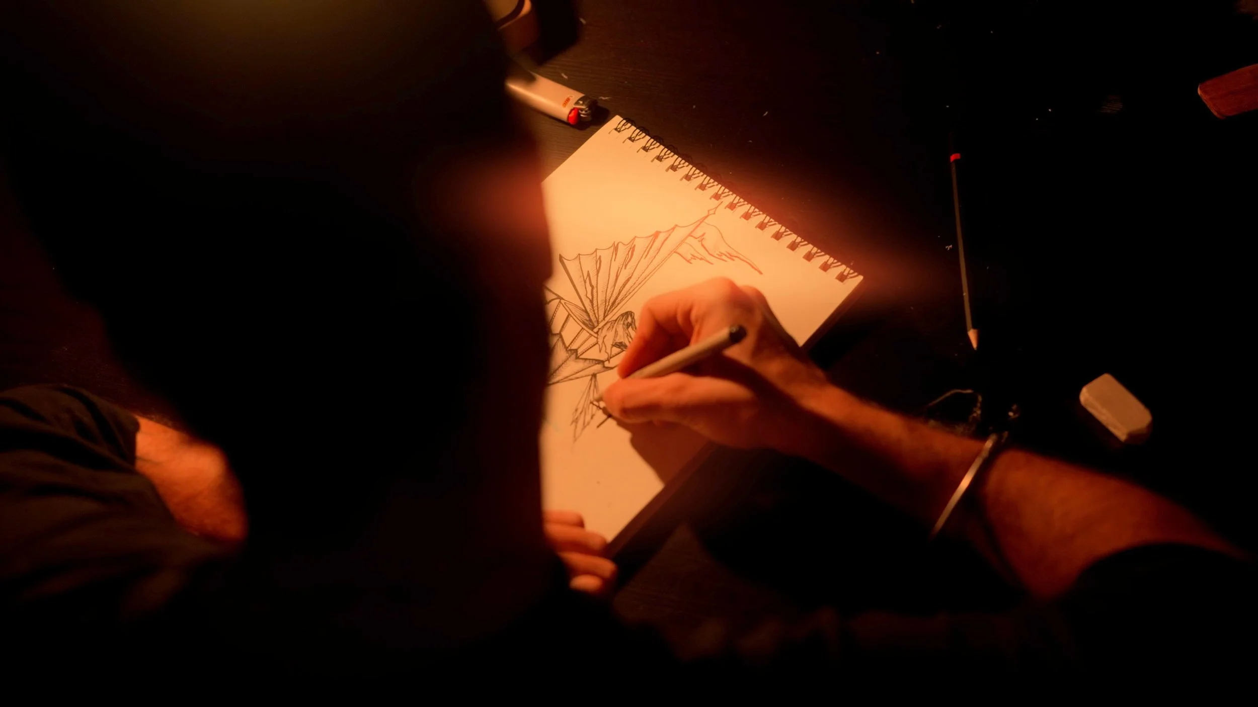 Une personne dessine un dragon dans un carnet à spirale, éclairée par une lumière chaude, avec un crayon à la main.