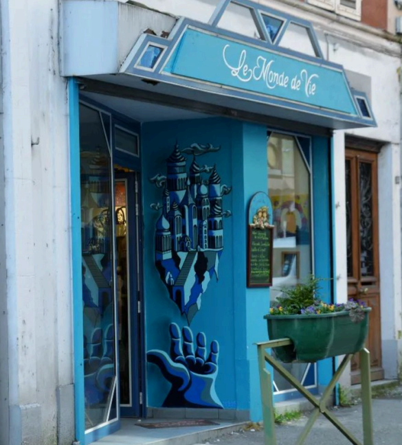 Façade d'une boutique appelée 'Le Monde de Vie' avec une fresque murale représentant une main tenant une ville avec des sapins et un ciel bleu.
