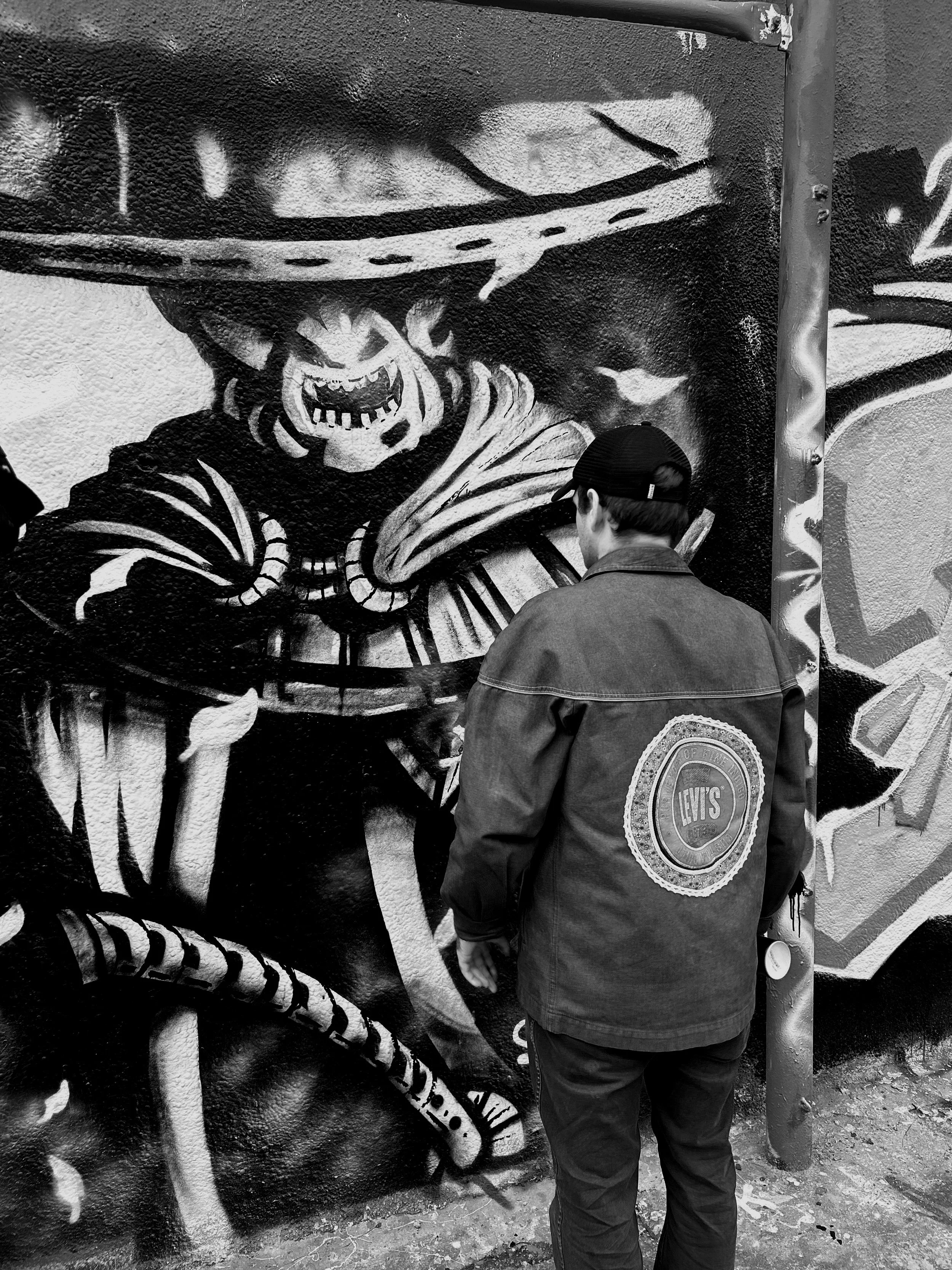 Un homme avec une veste portant un patch Levi's regarde une fresque murale en noir et blanc représentant un homme portant un chapeau et une écharpe, avec un visage effrayant.