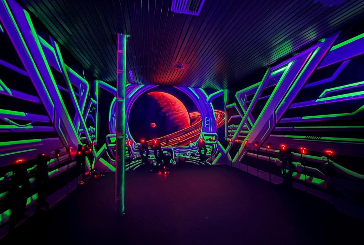 Salle d'attraction ou exposition futuriste avec une scène de planètes et d'anneaux dans un décor néon vert, violet et rouge, avec plusieurs personnes observant l'installation.