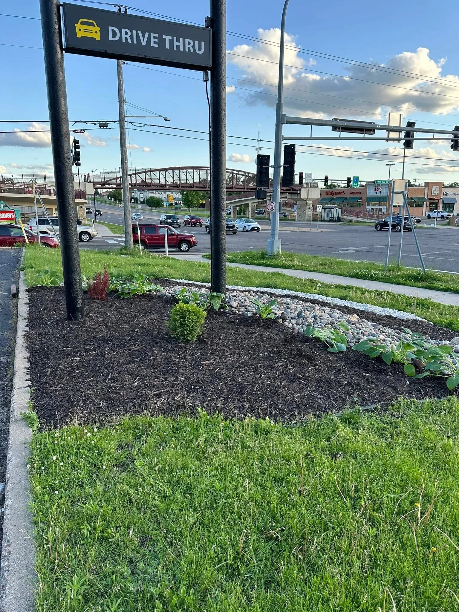 A parking lot with a small landscaped grassy area and a sign that reads 'Drive Thru' with a yellow car icon. The background shows a busy street with cars and commercial buildings including Walgreens and other stores, under a partly cloudy sky.