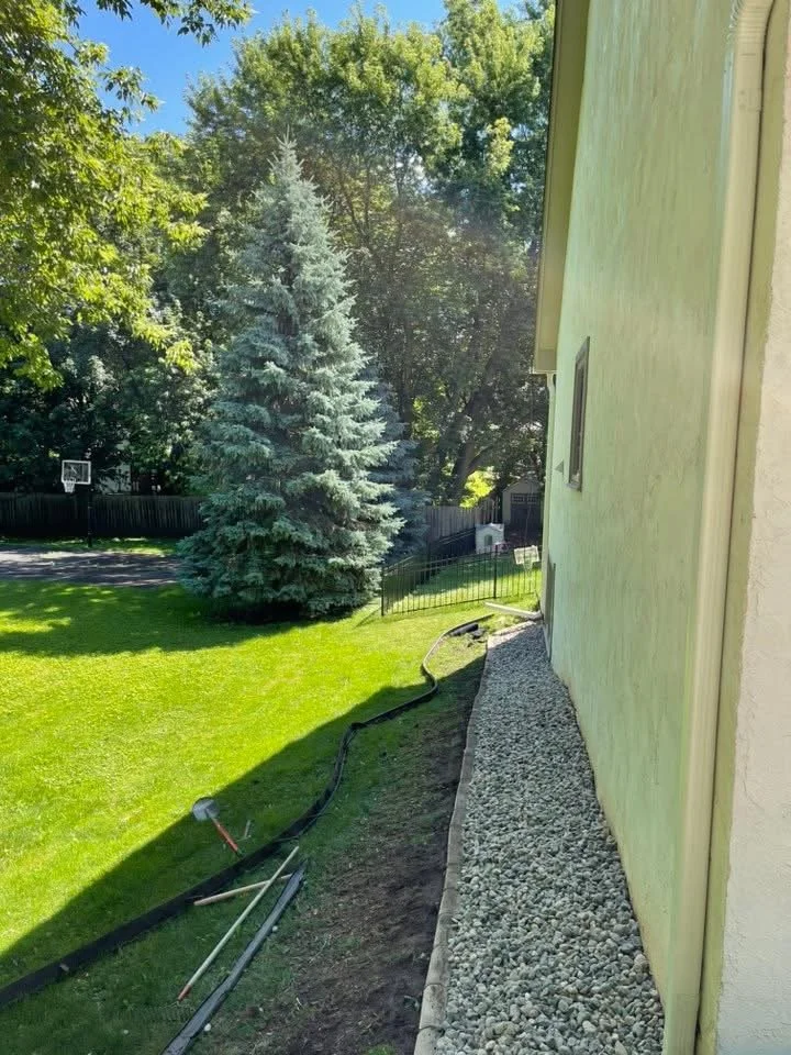 Backyard with green grass, tall evergreen tree, and surrounding trees in the background. Side of house with gravel pathway and garden hose.