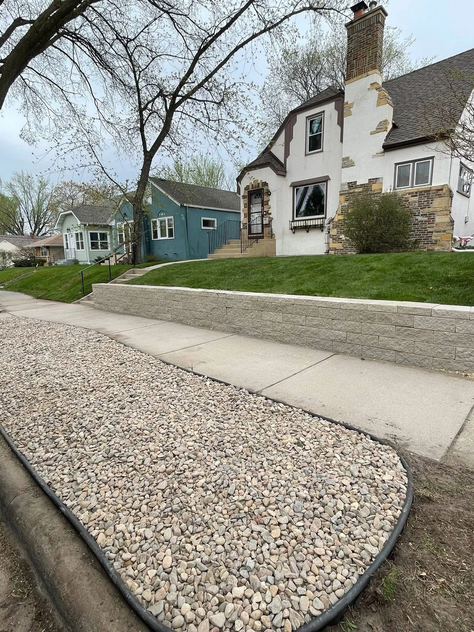 A sidewalk runs along a hillside with a retaining wall, leading to houses with yards. The houses are painted in white and blue, with some brick and stone accents. There are trees with budding leaves and an overcast sky.