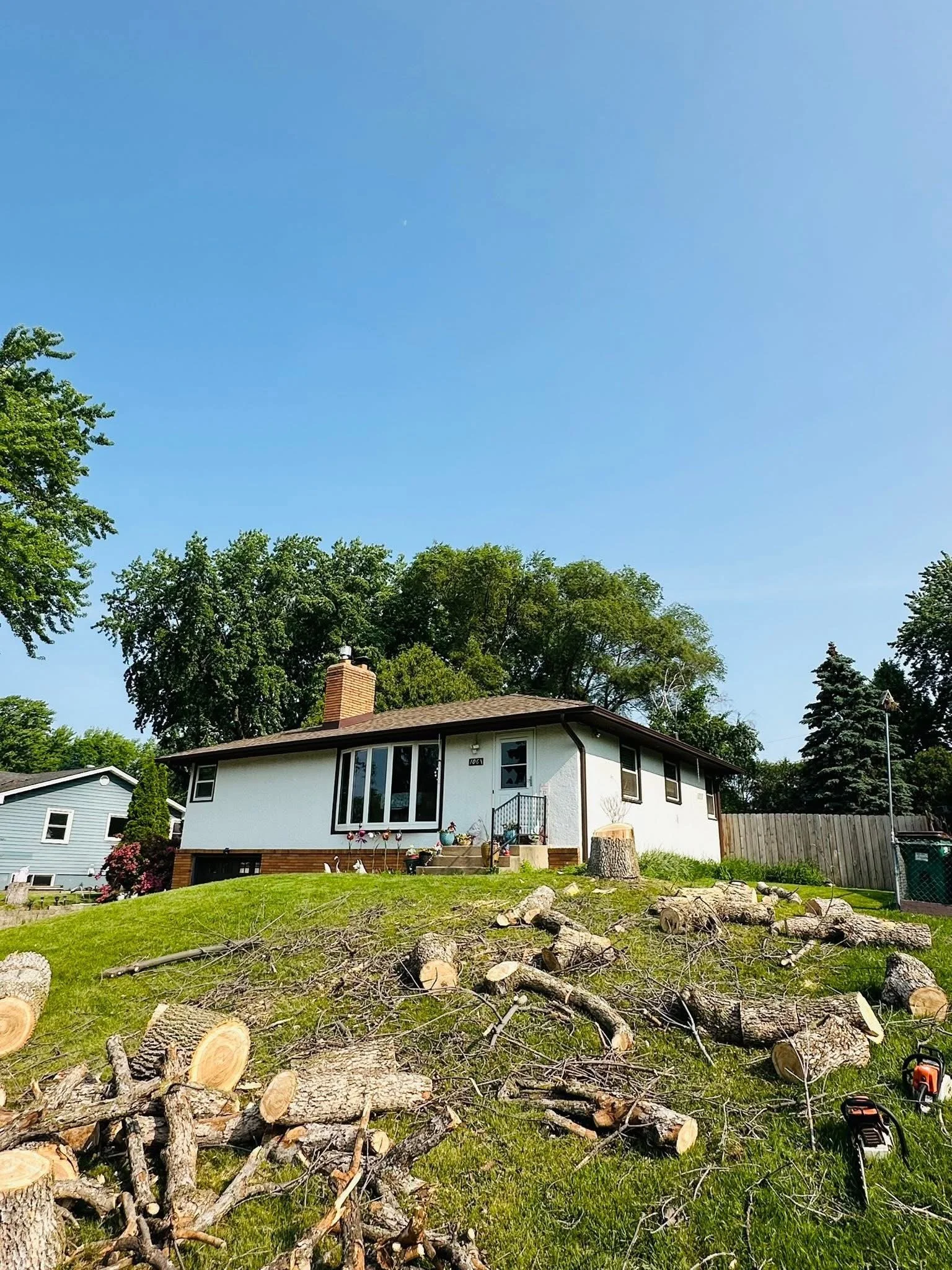 A house with a brick chimney and large front window, surrounded by trees and a grassy yard with cut tree logs and branches, and two chainsaws on the ground.