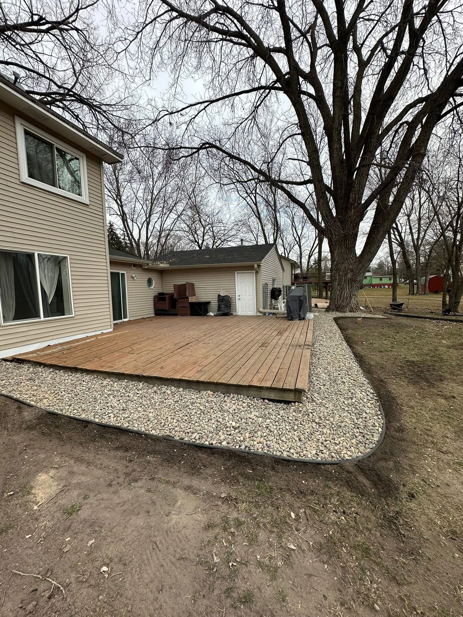 A backyard with a new wooden deck, gravel border, and large tree, neighboring houses visible in the background.