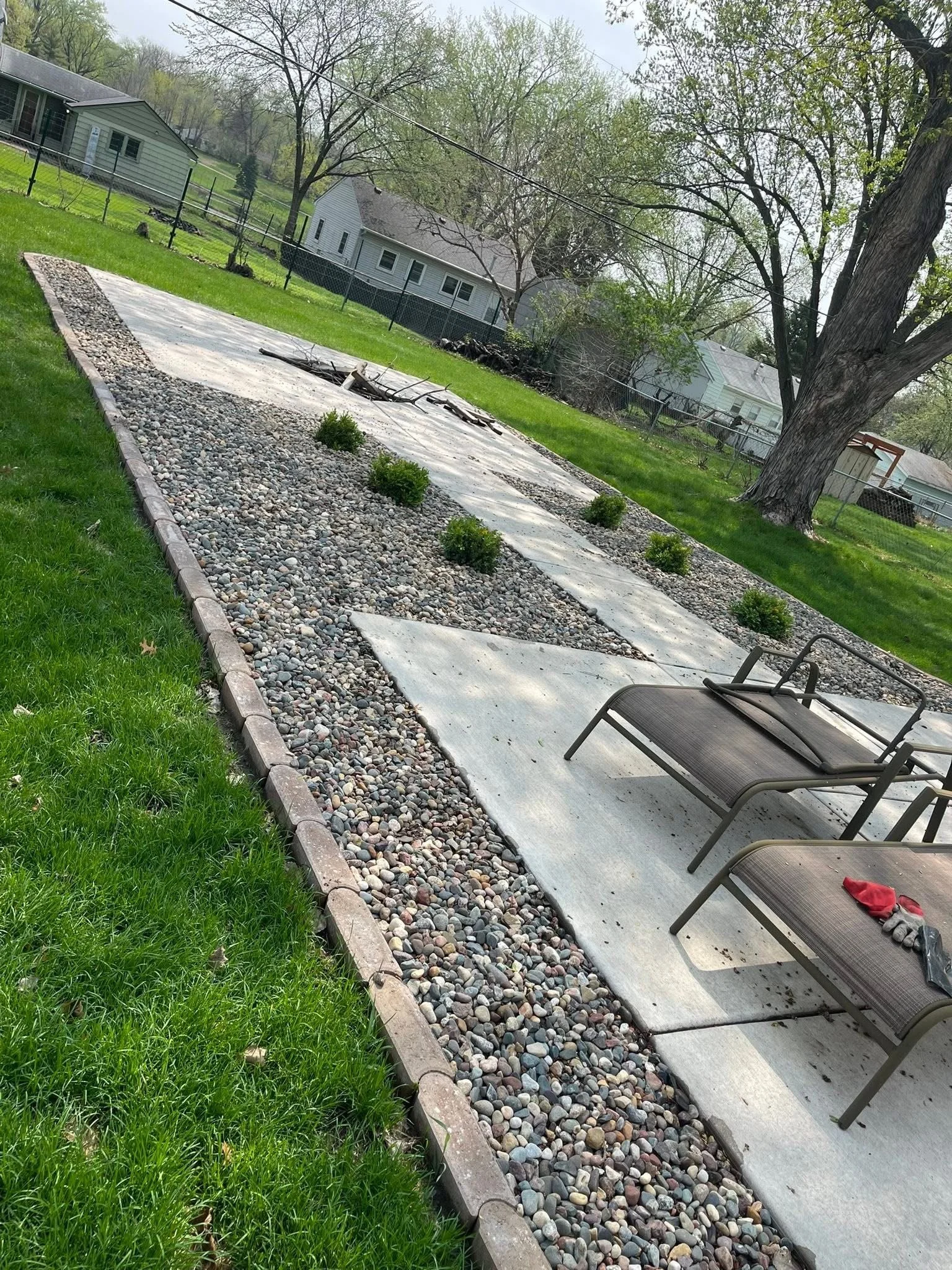 Backyard with a concrete patio, two lounge chairs, small shrubs, rocky garden bed bordered by bricks, large tree, and neighboring houses in the background.