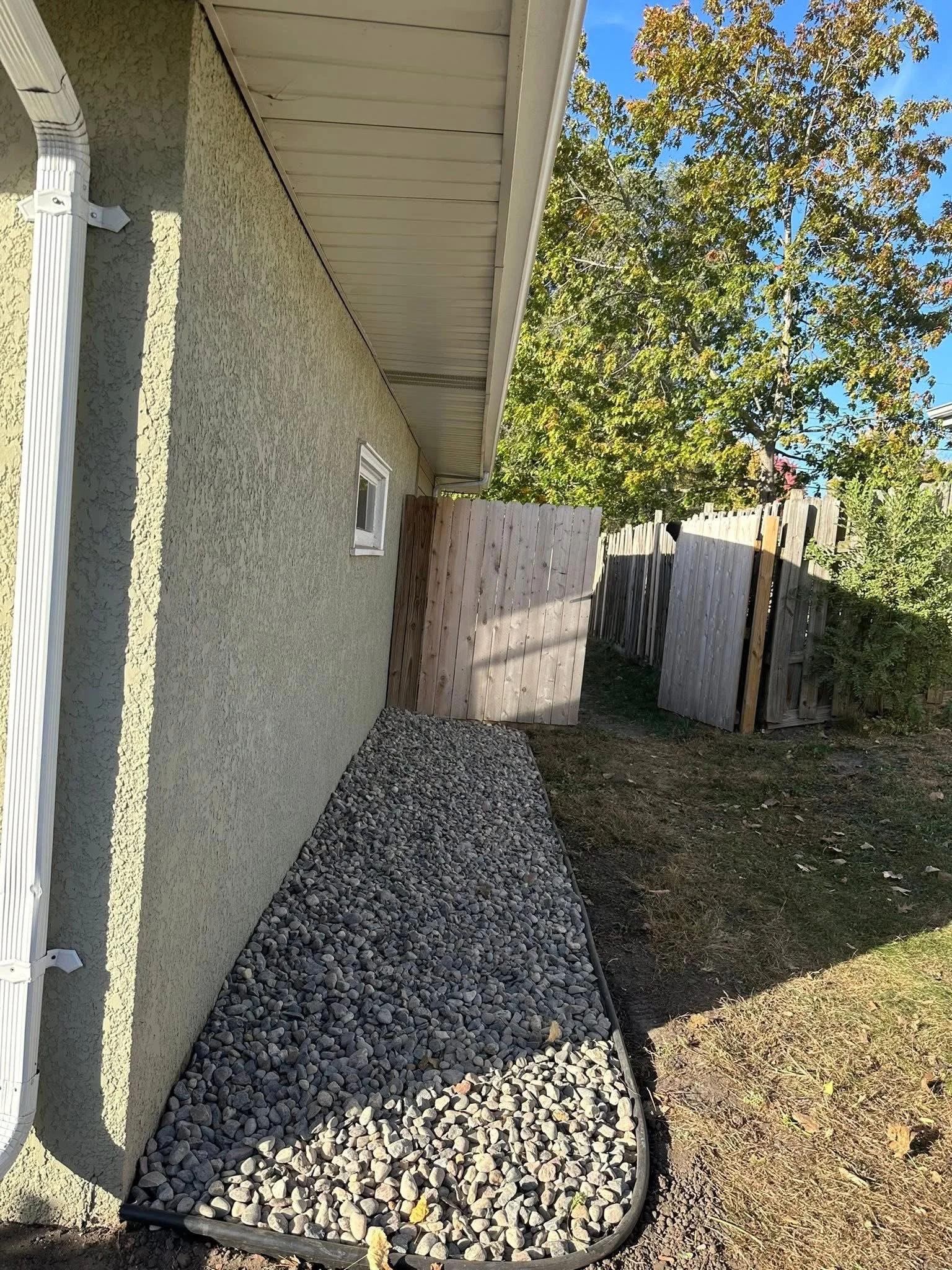 Side yard with gravel landscaping, a green stucco house wall, a small window, a wooden fence and a large tree with green leaves