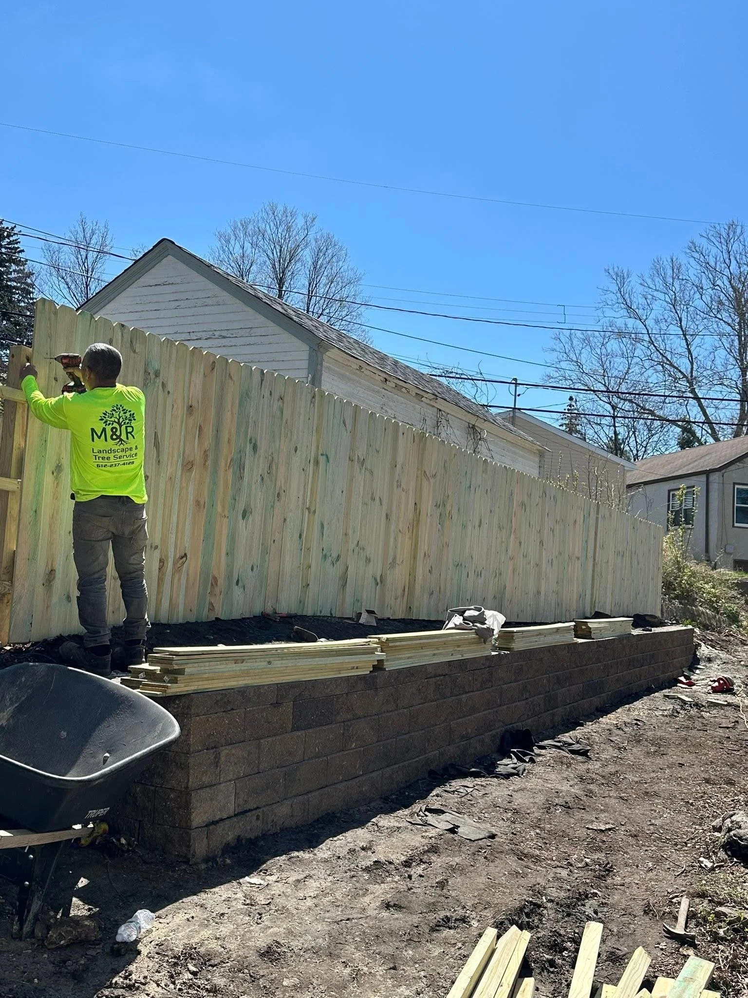 A person in a bright neon yellow shirt working on building a wooden fence in a backyard, with a house and trees in the background.