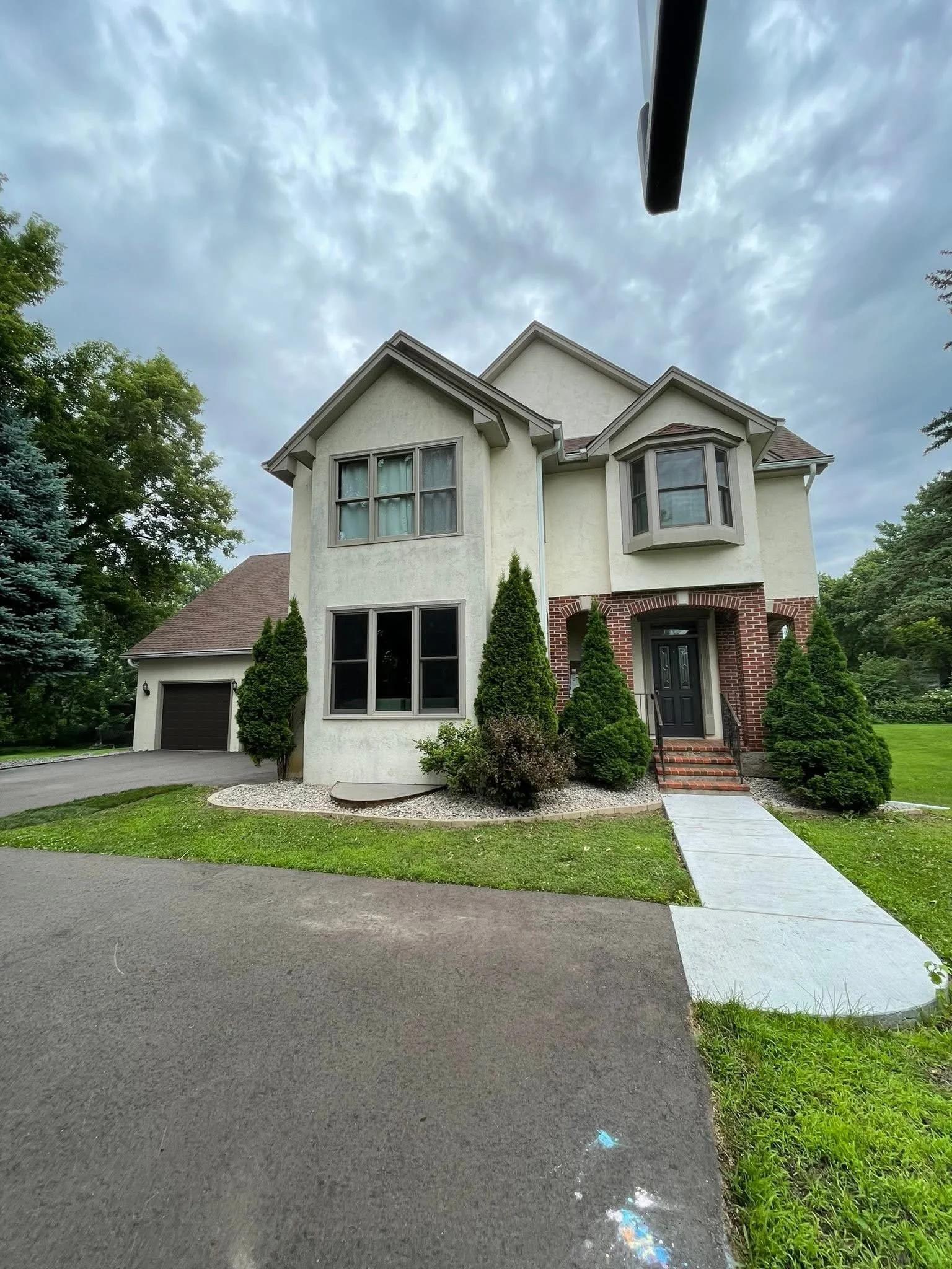A two-story house with a white exterior, brick accents at the entrance, and a dark front door. The house is surrounded by green trees and bushes. There is a concrete walkway leading to the front door and a driveway with an attached garage on the left