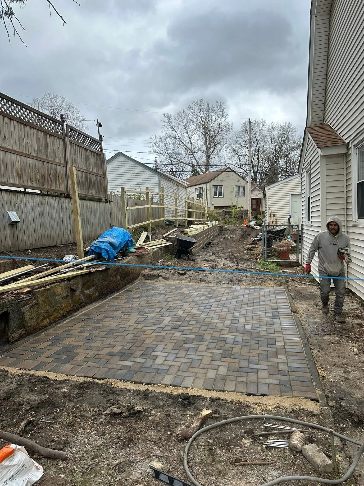 A person with a hoodie and gloves walking on a backyard patio under construction, with a recently laid paved brick surface. There are construction materials, a wheelbarrow, and tools scattered around. The yard is fenced, with trees and neighboring ho