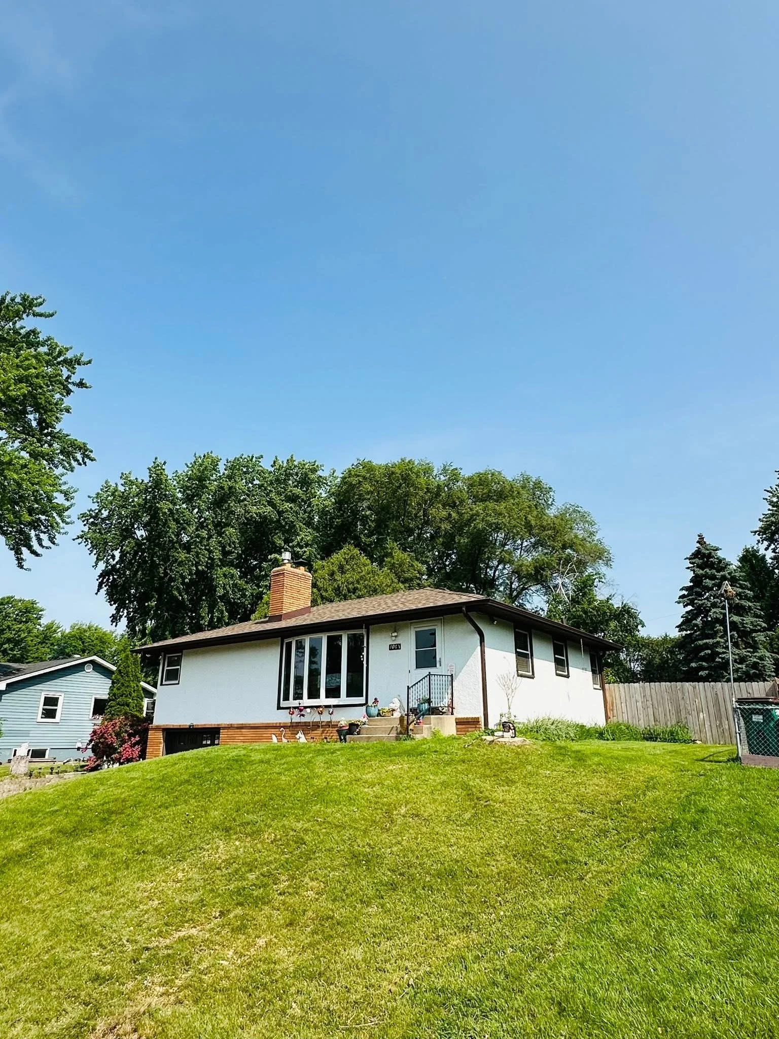 A white house with a brick chimney and large front window, surrounded by a well-maintained grassy yard with decorative items on the steps, under a clear blue sky with trees in the background.