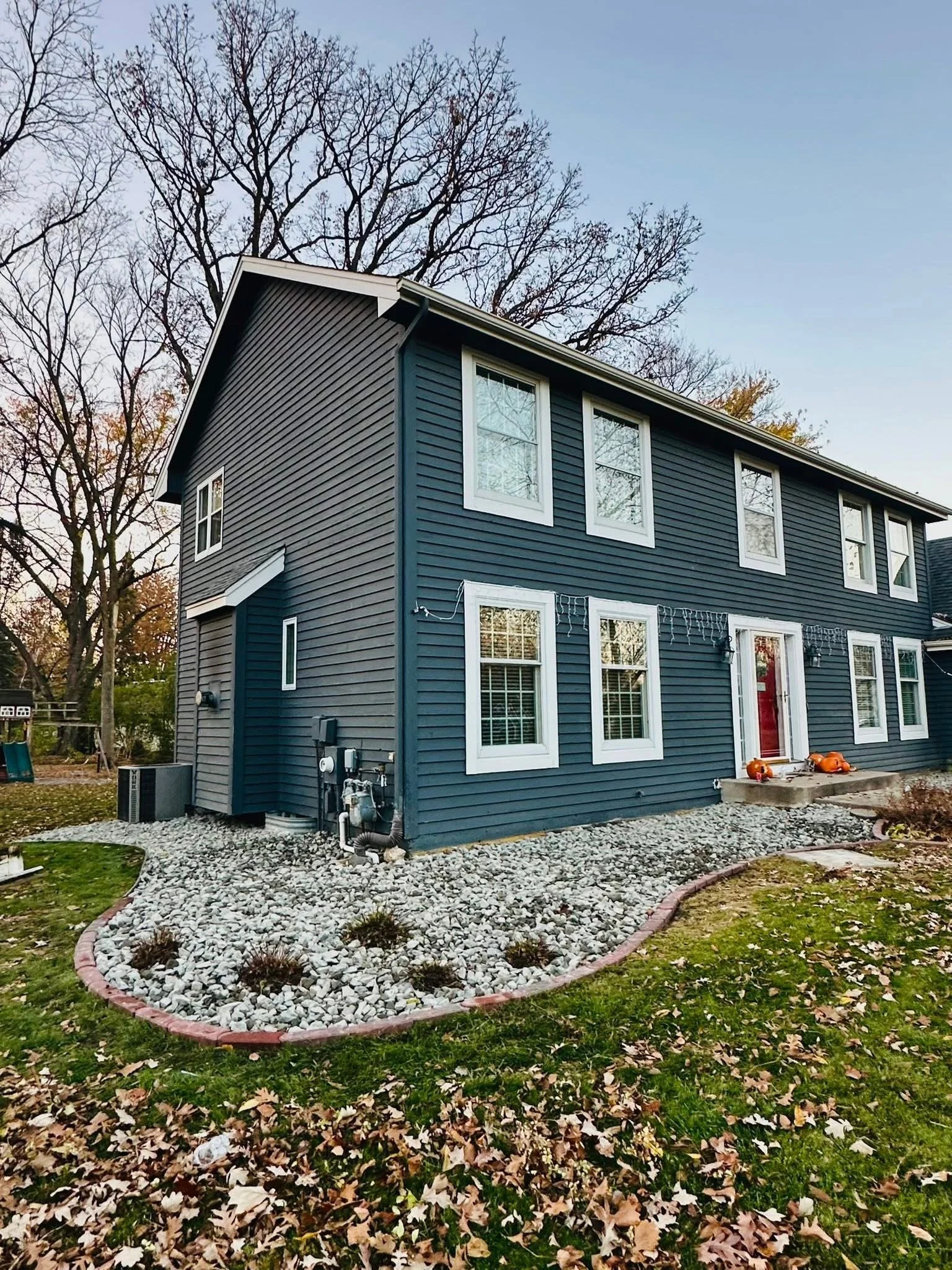Blue two-story house with white-framed windows, red front door, and decorated pumpkins outside, surrounded by leaf-covered ground and a tree in the background.