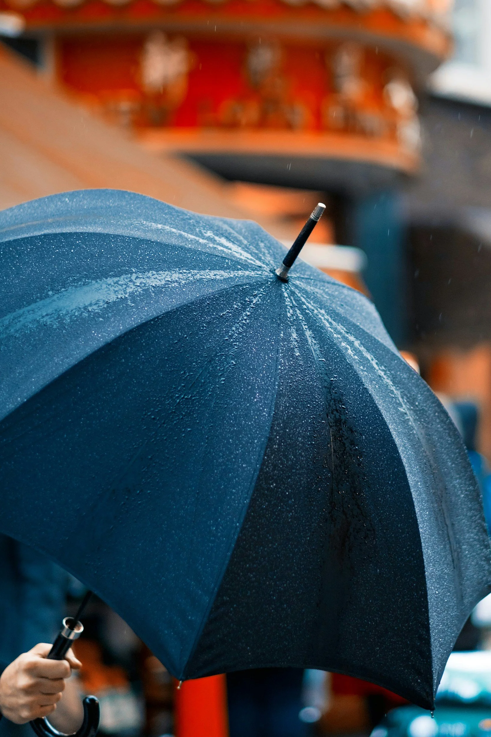 Close-up of a blue umbrella with raindrops on it, held by a person with a black raincoat, with a blurred background of a building and people in an outdoor setting.