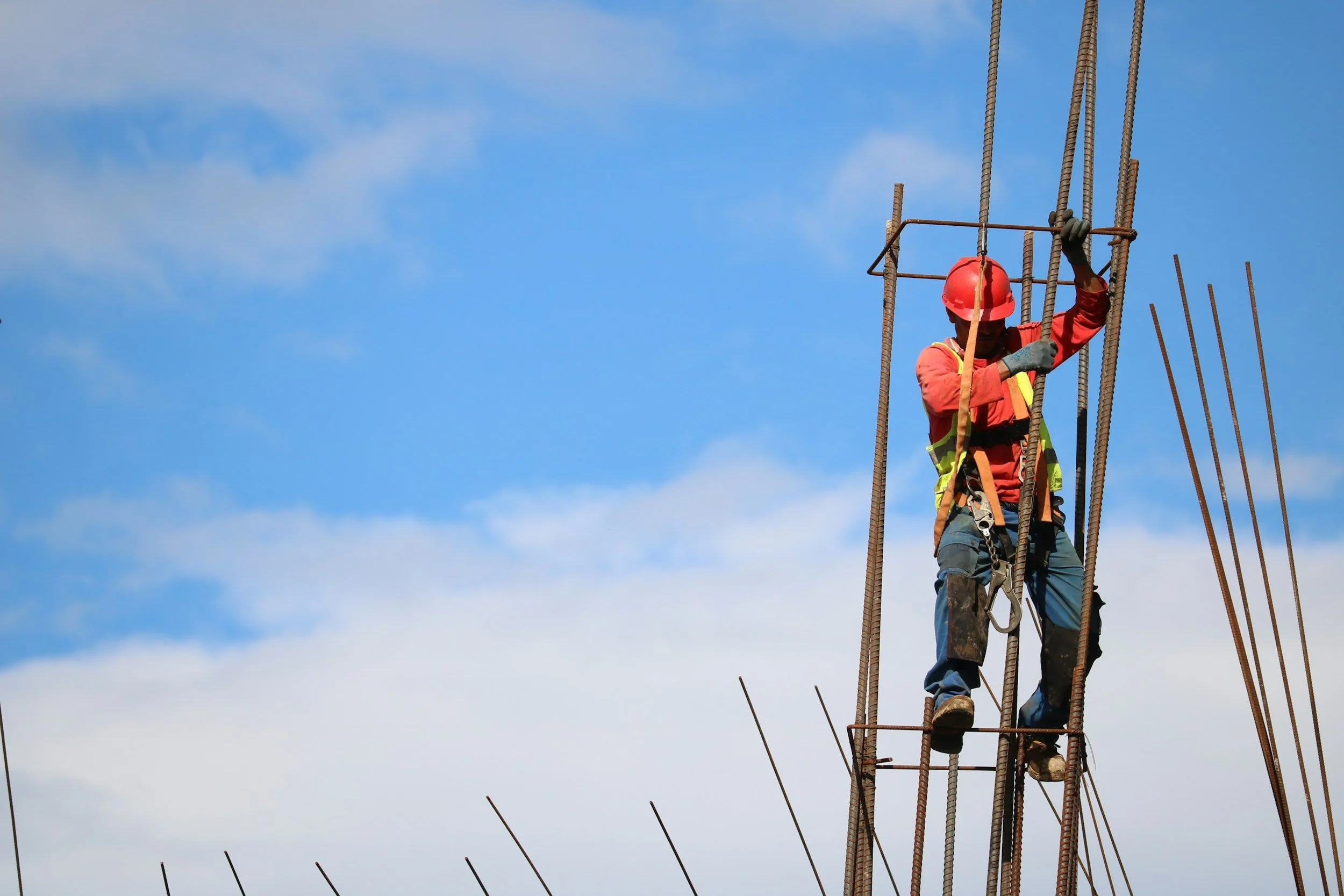 Construction worker wearing a red safety helmet, orange jacket, and safety harness working among vertical steel reinforcement bars at a construction site under a blue sky.
