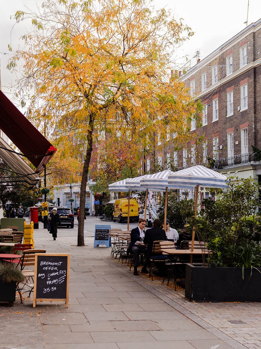 City street with outdoor cafe, striped umbrellas, people sitting and talking, leafless trees with autumn leaves, parked cars, a yellow delivery van, and a cloudy sky.