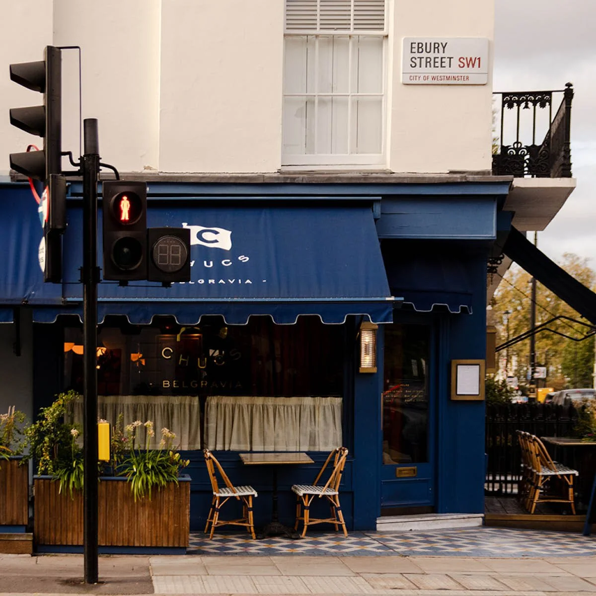 Street view of a restaurant with dark blue facade, outdoor seating, and a traffic light showing red pedestrian signal at Ebury Street in Westminster. The restaurant has a blue awning with white lettering.