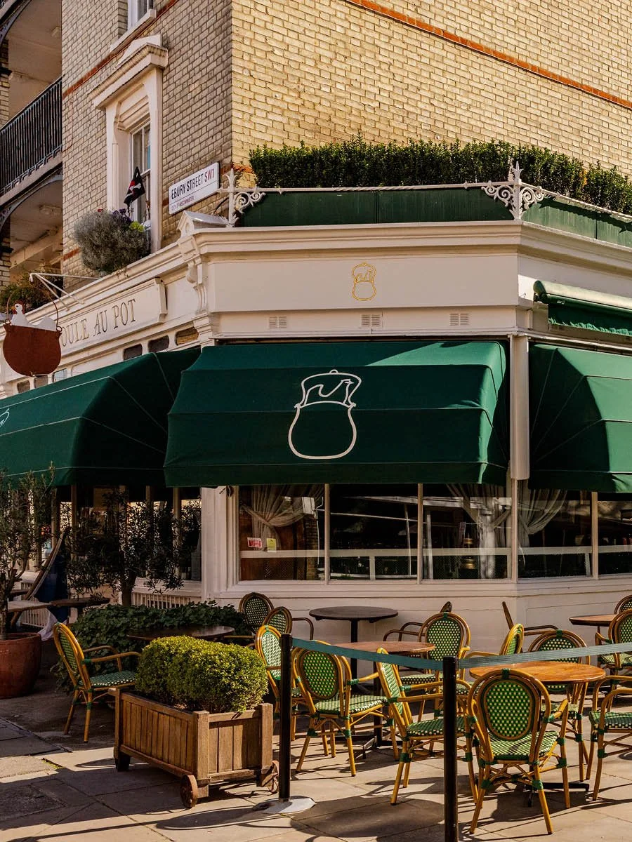 Outdoor seating area of a café with green umbrellas, yellow chairs with green cushions, and potted plants, on a city street corner in front of a building with yellow brick walls and a white façade.