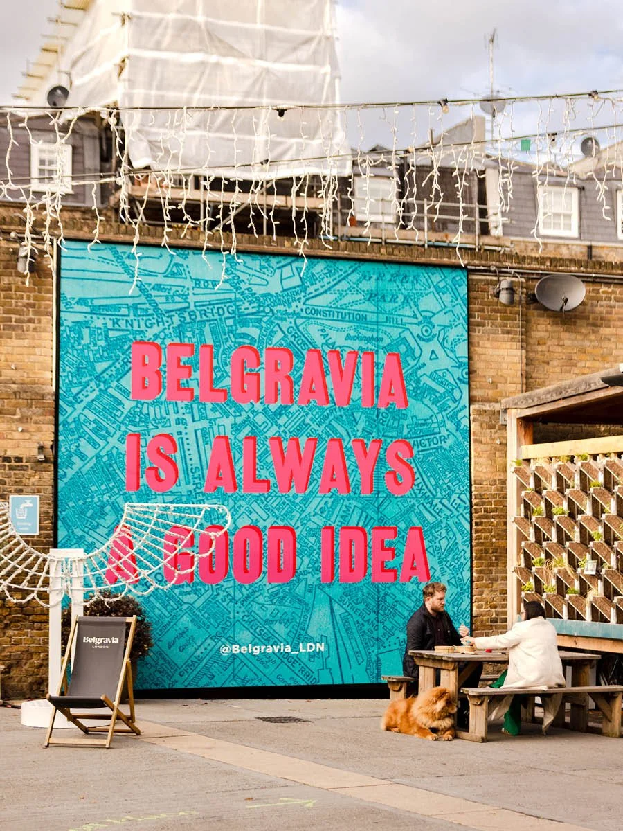 Large colorful sign with a city map background reads "Belgravia is Always a Good Idea" at an outdoor cafe with two people sitting at a picnic table and a fluffy dog nearby. String lights hang above.