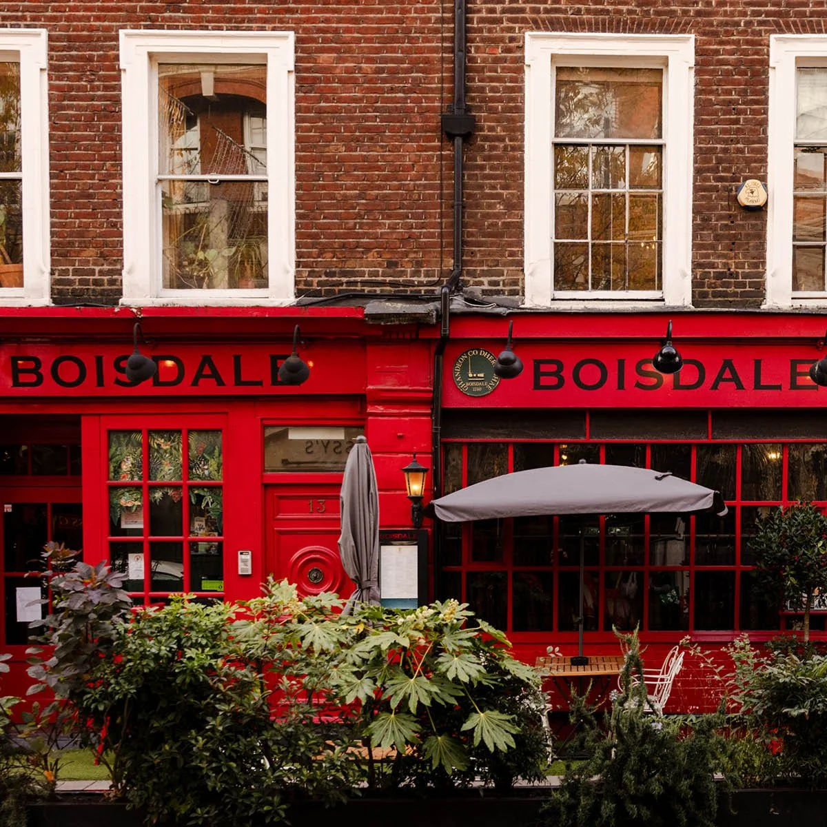 Red storefront with black sign that reads 'BOISDALE', large windows, outdoor seating area with plants, umbrella, and a lantern, brick building above with three white-framed windows.