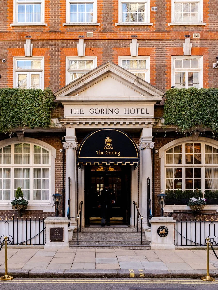 Exterior of The Goring Hotel, a historic black and white brick building with white-framed windows, ivy on the facade, and a black awning with gold lettering and crest at the entrance, with a person in a dark suit standing under the awning.