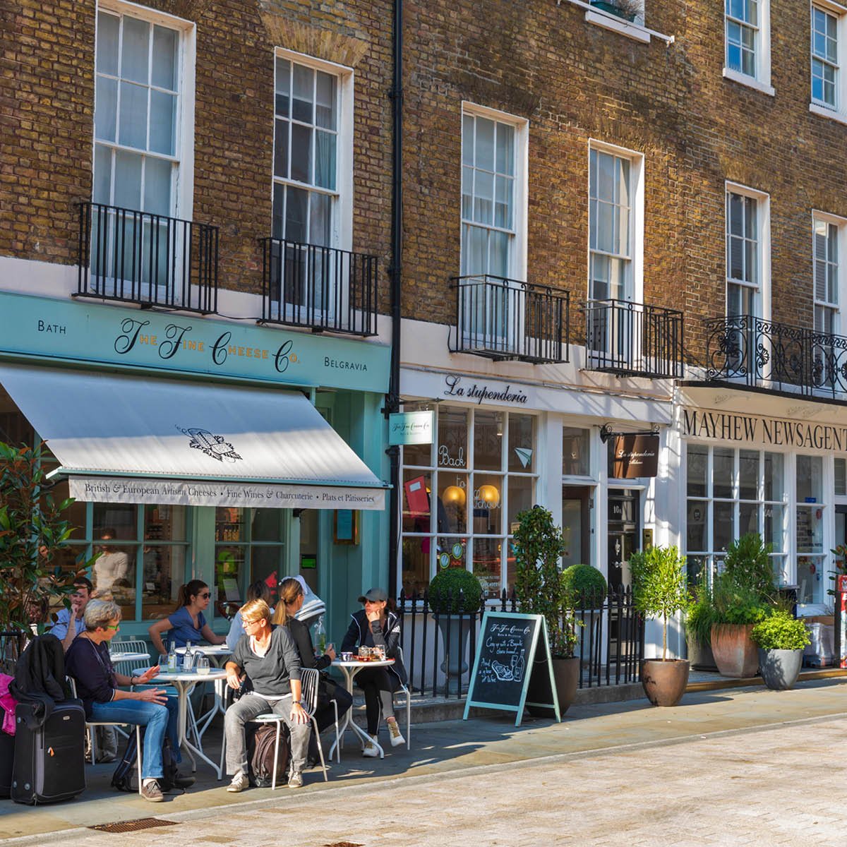 People sitting and chatting outside cafes on a sunny street, with storefronts and potted plants along a brick building.