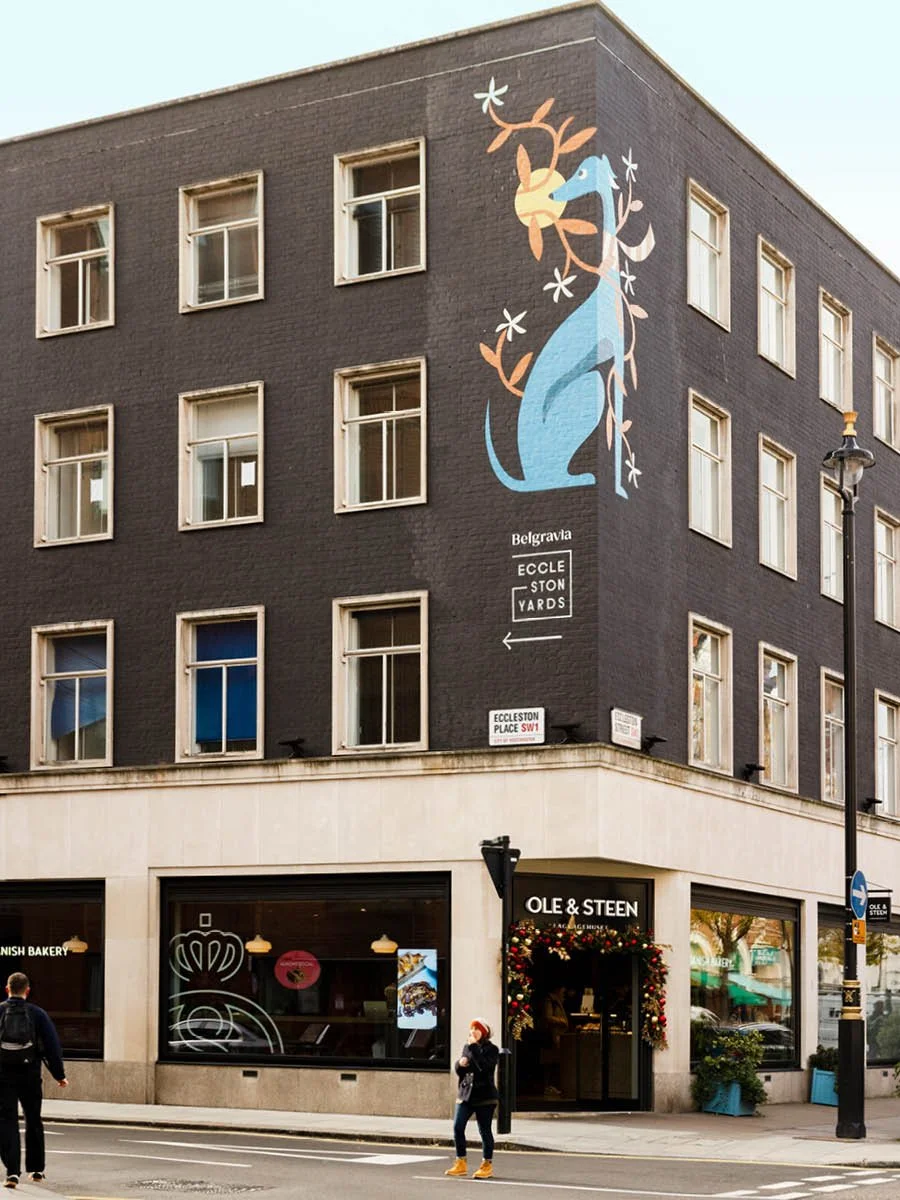 Street corner with building featuring a large mural of a blue dinosaur intertwined with orange branches and white flowers. The building has multiple windows and a shop called Ole & Steen on the ground floor, decorated for the holidays.