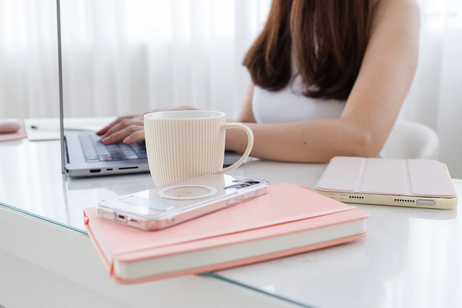 A mug, notebook and cellphone, with website designer in the background, working on a website audit on her laptop