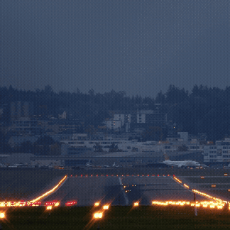 Nighttime view of an airport runway with illuminated lights guiding aircraft takeoff and landing, with buildings and trees in the background