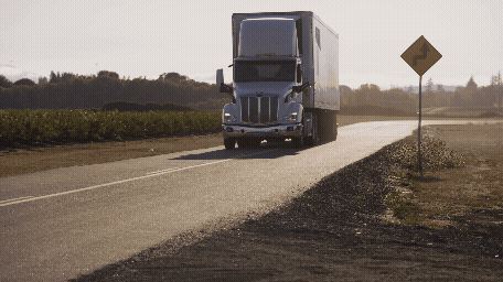 A semi-truck driving down a rural road with fields and trees in the background.