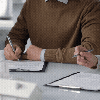 Person in a brown sweater writing with a pen on a clipboard at a desk.