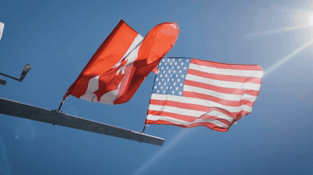 Canadian and American flags flying in the wind against a clear blue sky.