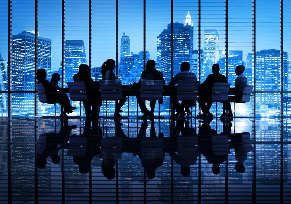 Silhouettes of nine people sitting at a conference table with a city skyline view through large windows