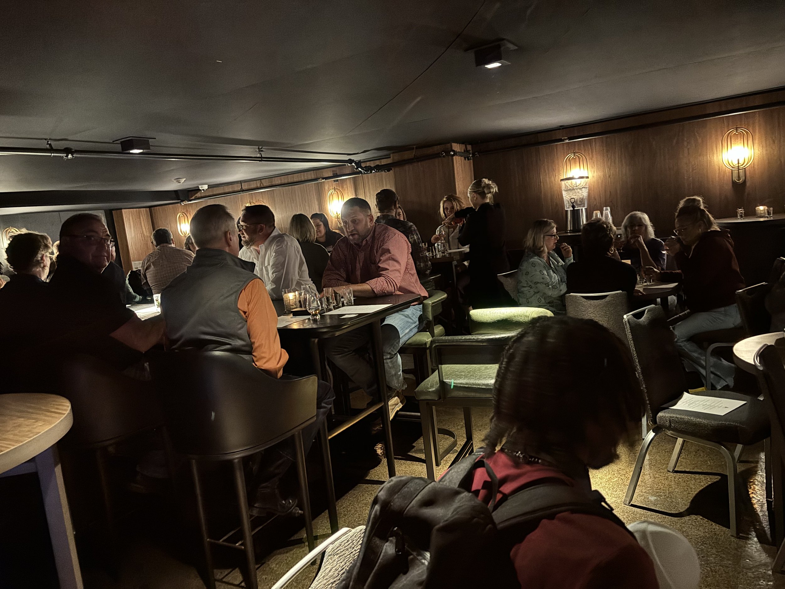 A dimly lit bar with people seated at tables and barstools, engaged in conversation. The atmosphere is cozy with small hanging lights and wooden walls. Several tables have drinks, and some patrons are wearing casual attire.