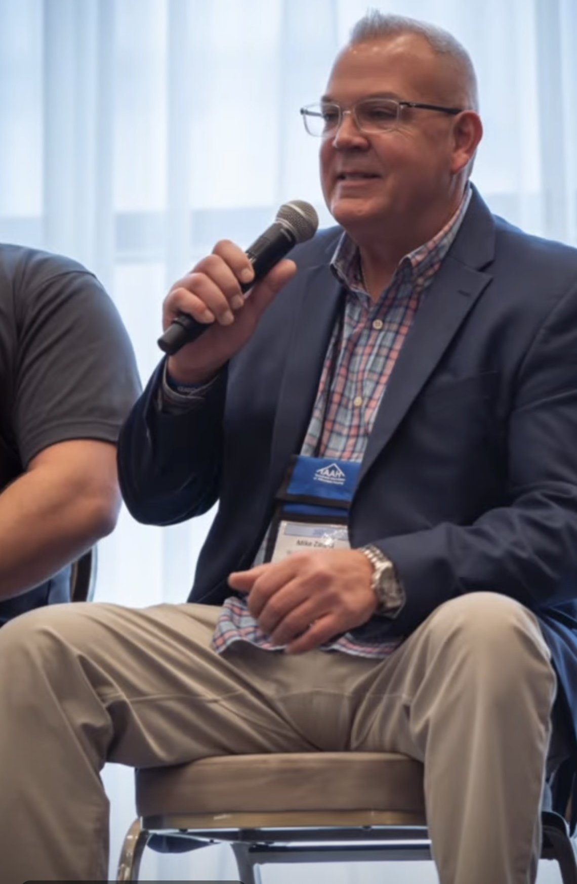 A man wearing glasses, a blue blazer, and a plaid shirt is sitting on a chair, holding a microphone and speaking. He has a conference badge around his neck.