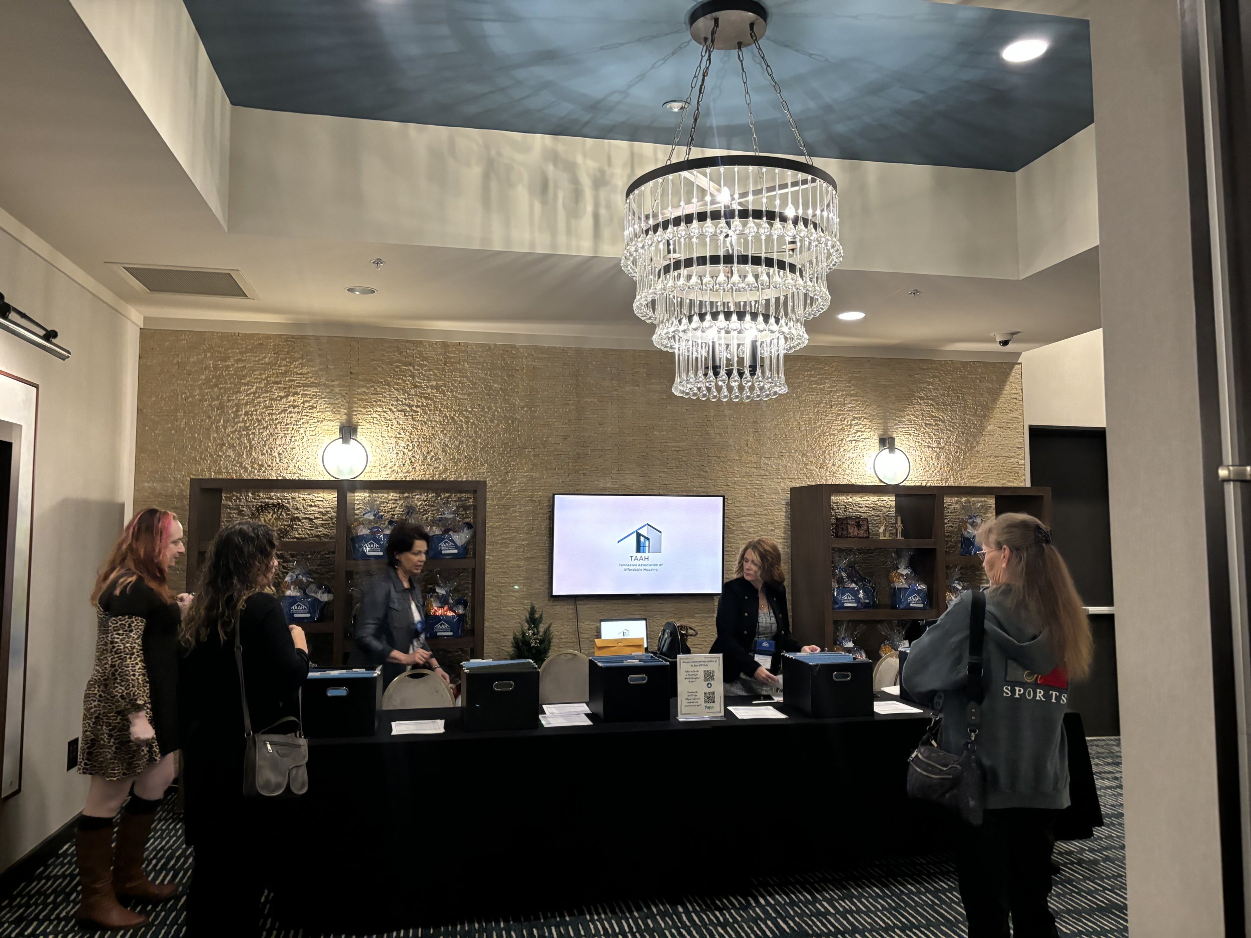 People interacting at a registration or information desk in a well-lit room with a chandelier and decorative wall. The desk has boxes, papers, and a sign with a QR code. Shelves in the background hold promotional bags with a logo.